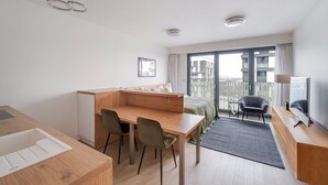 Ein modernes Hotelzimmer mit Schreibtisch aus Holz, zwei Stühlen, einem Bett mit grüner Bettdecke, einem kleinen Balkon mit einem Stuhl und einem großen Fenster mit Blick auf die Stadt.