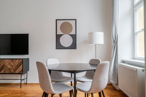 Uma sala de estar moderna com uma mesa redonda, cadeiras estofadas cinzentas, uma televisão de tela plana, um armário de madeira, um abajur e um radiador.