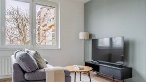 Ein modernes Wohnzimmer mit einem grauen Sofa, einem runden Couchtisch, einem Fernseher auf einem schwarzen Standfuß und einem Fenster mit Blick auf Gebäude und kahle Bäume.