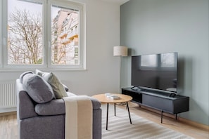 Un salón moderno con un sofá gris, una mesa de centro redonda, un televisor sobre un soporte negro y un ventanal con vistas a edificios y árboles desnudos.