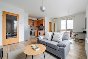Un apartamento moderno con sofá gris, suelo de madera, cocina con armarios de madera, zona de comedor y un ventanal con vistas.