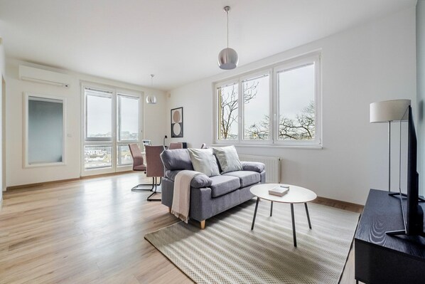 Un salon spacieux avec un canapé gris, une table basse ronde et un meuble pour la télévision. De grandes fenêtres laissent entrer la lumière naturelle et offrent une vue sur la ville.