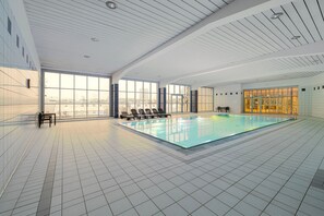 Une piscine intérieure moderne avec des parois en verre transparent, un sol carrelé et une vue sur le paysage urbain.
