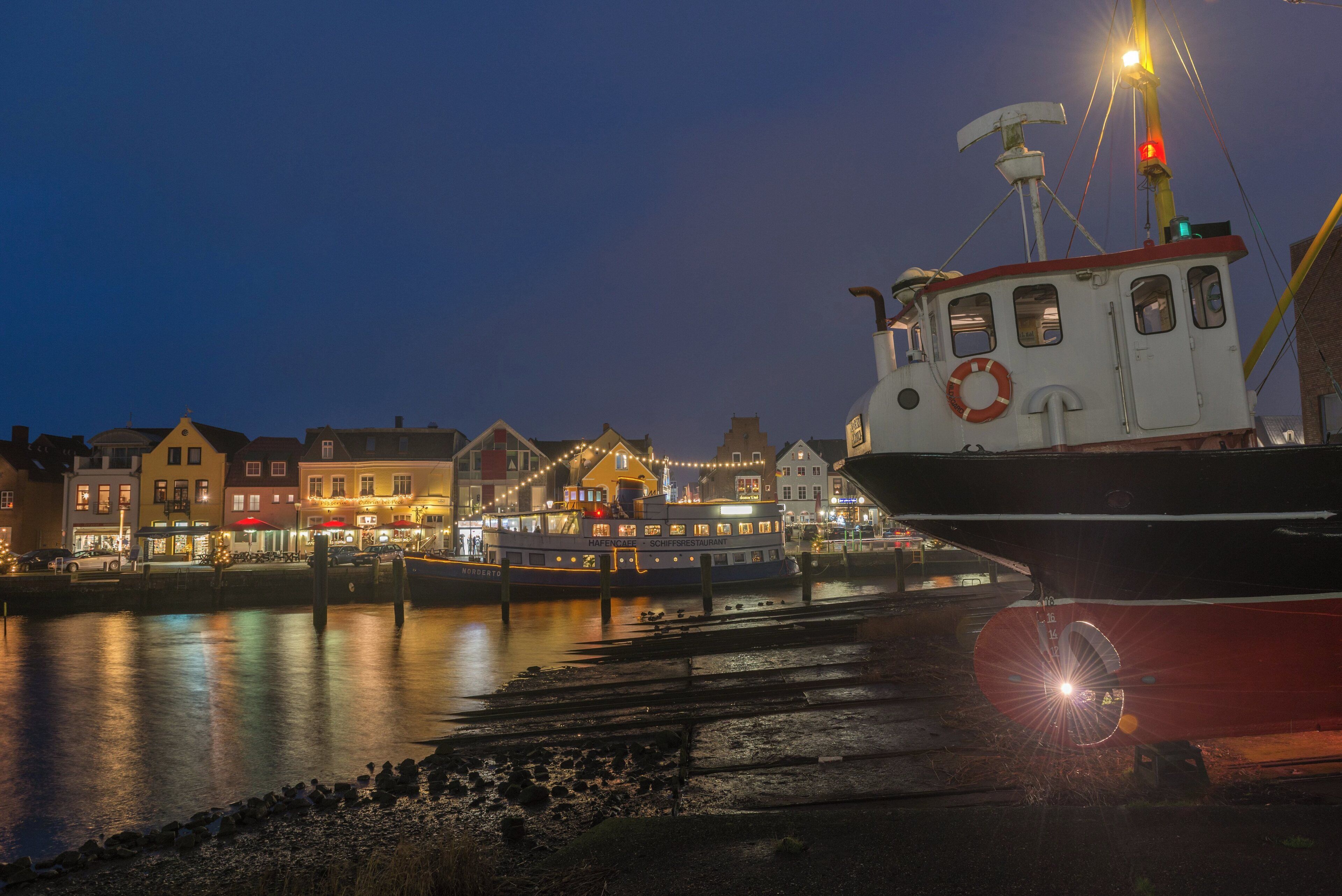 Une scène au bord de l’eau, avec un bateau amarré près d’immeubles dont les fenêtres sont illuminées, ce qui laisse supposer une ambiance urbaine animée.