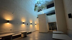 Ein modernes Interieur mit Bänken und Wandleuchten, einer großen zentralen Säule und Blick auf einen Baum durch eine Glastür.