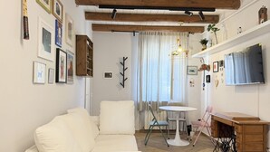 Ett vardagsrum med en vit soffa, ett litet runt bord, ett skrivbord i trä och en tv på väggen.