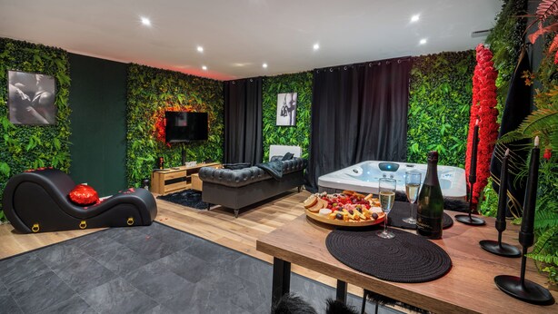 A modern living room with a green wall, a black sofa, a wooden table with a tray of snacks, a hot tub, and a TV mounted on the wall.