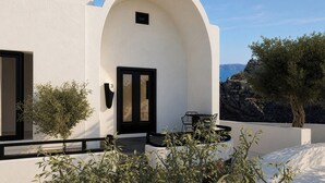 A modern building with a white facade, a black door, and a small window. There is a table and chairs outside, and a tree is visible in the background. - Mamay Hotel (Santorini)