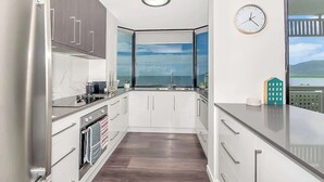 A modern kitchen with a view of the sea, white cabinets, stainless steel appliances, and a clock on the wall. - Luxury Waterfront: Spectacular Oasis in the City (Cairns North)