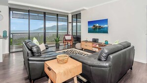 A modern living room with a black leather sofa, a wooden coffee table, and a large window offering a view of the sea. - Luxury Waterfront: Spectacular Oasis in the City (Cairns North)