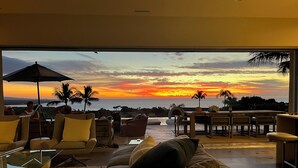 Amplia sala de estar con un gran ventanal que ofrece vistas al atardecer, amueblada con sofás, sillas y una mesa de comedor.
