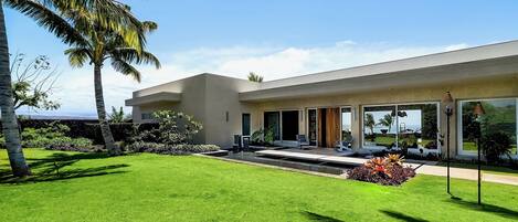A modern house with a well-maintained lawn, palm trees, and a clear view of the ocean.