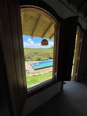 A room with a large window overlooking a swimming pool and a grassy landscape. - Tupinambá Hostel Caraíva (Porto Seguro)