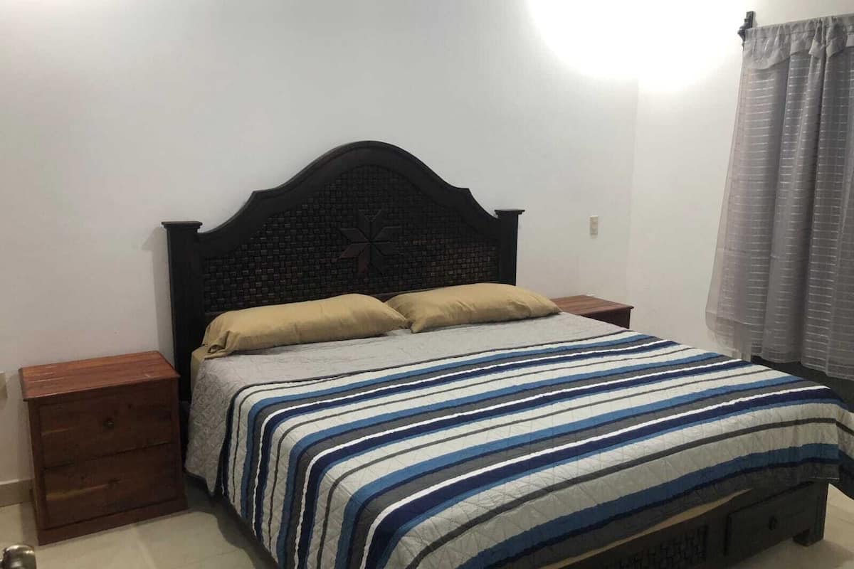 A bedroom with a dark wooden headboard, a striped bedspread, two pillows, a nightstand, and a window with curtains.