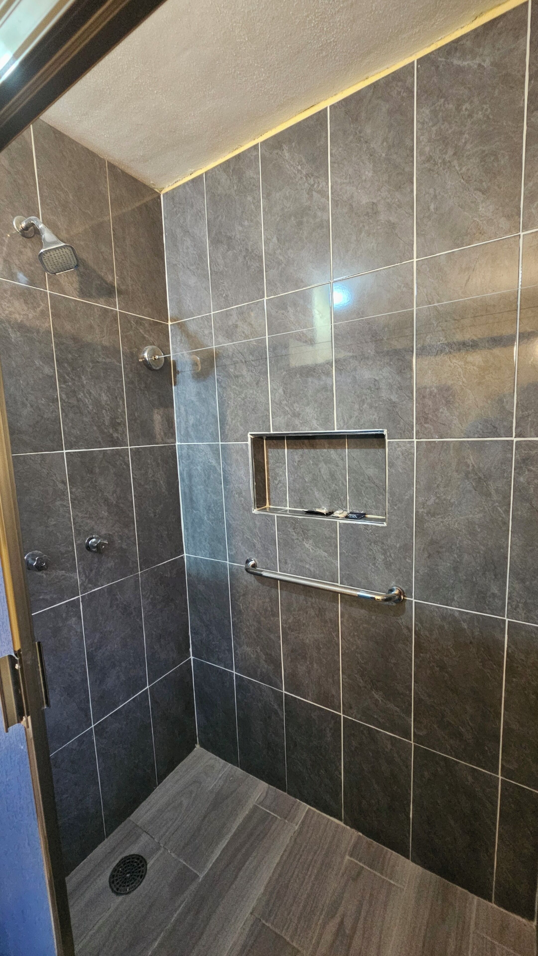 Una ducha a nivel con azulejos oscuros, un estante metálico y un cabezal de ducha manual.