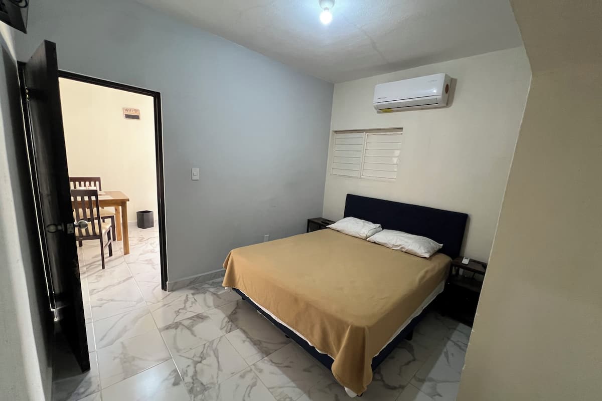 A bedroom with a bed, a nightstand, a wall-mounted air conditioner, and a door leading to a dining area.