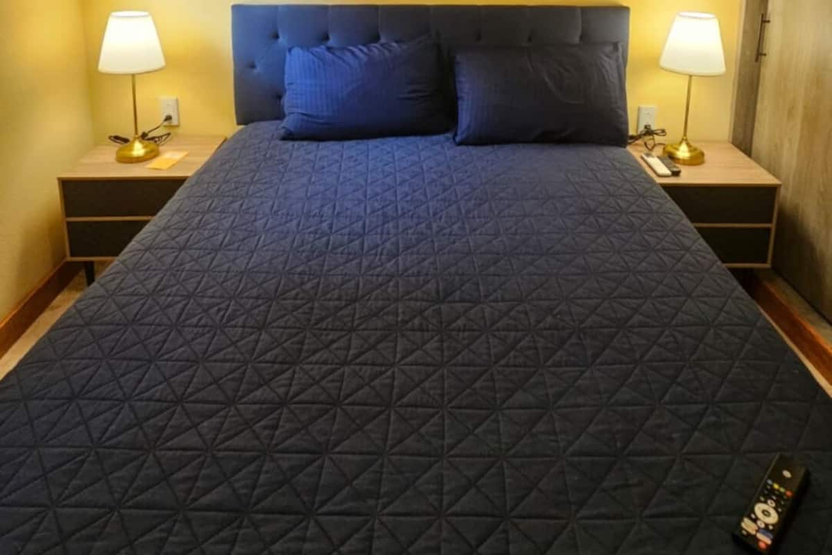 A bed with a geometric quilt, two bedside lamps, a painting of a night sky, and a remote control on the bed.