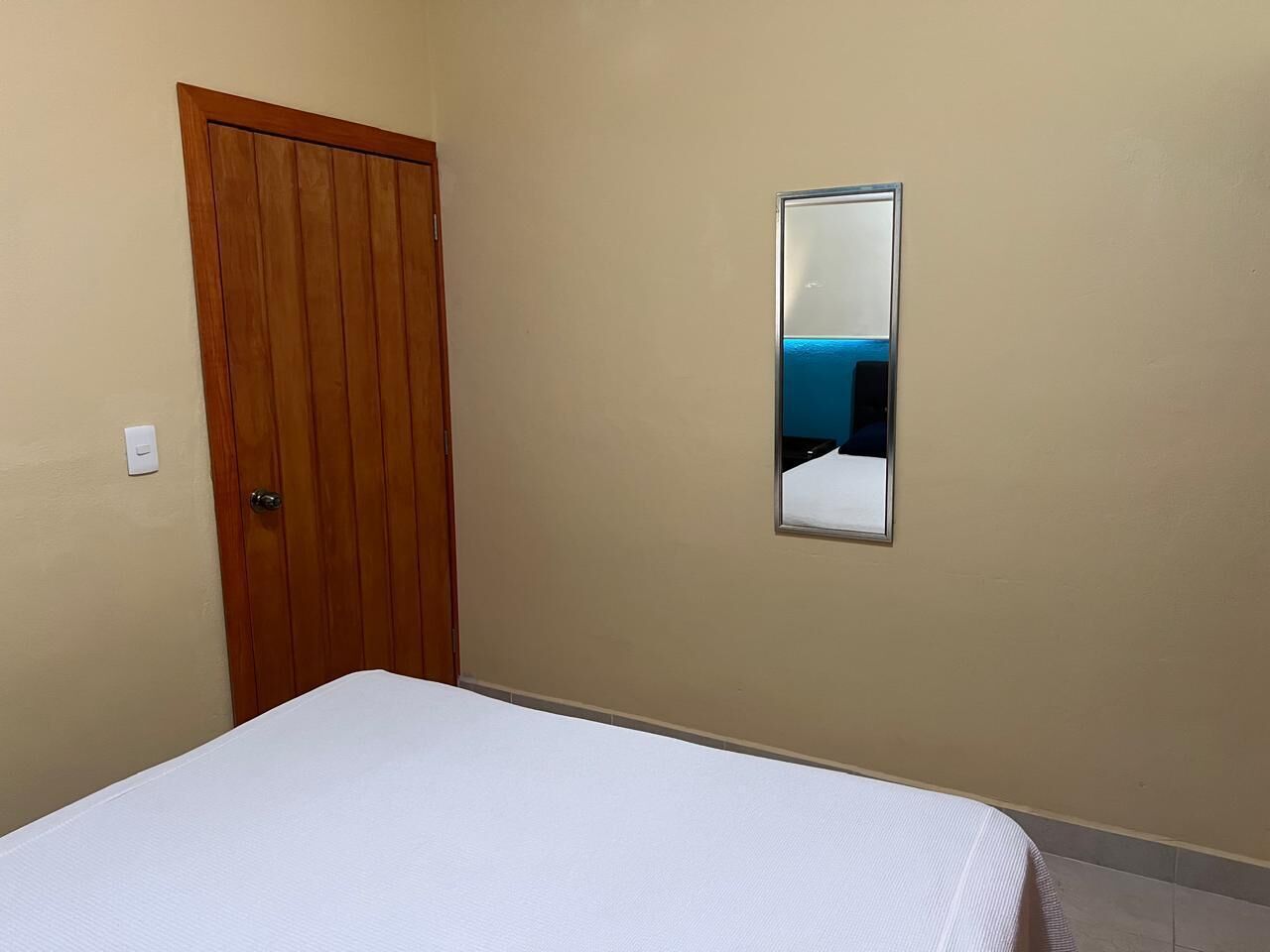 Un dormitorio con puerta de madera, cama blanca y espejo montado en la pared.