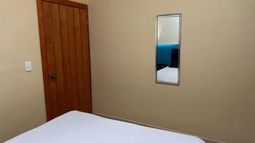 Un dormitorio con puerta de madera, cama blanca y espejo montado en la pared.