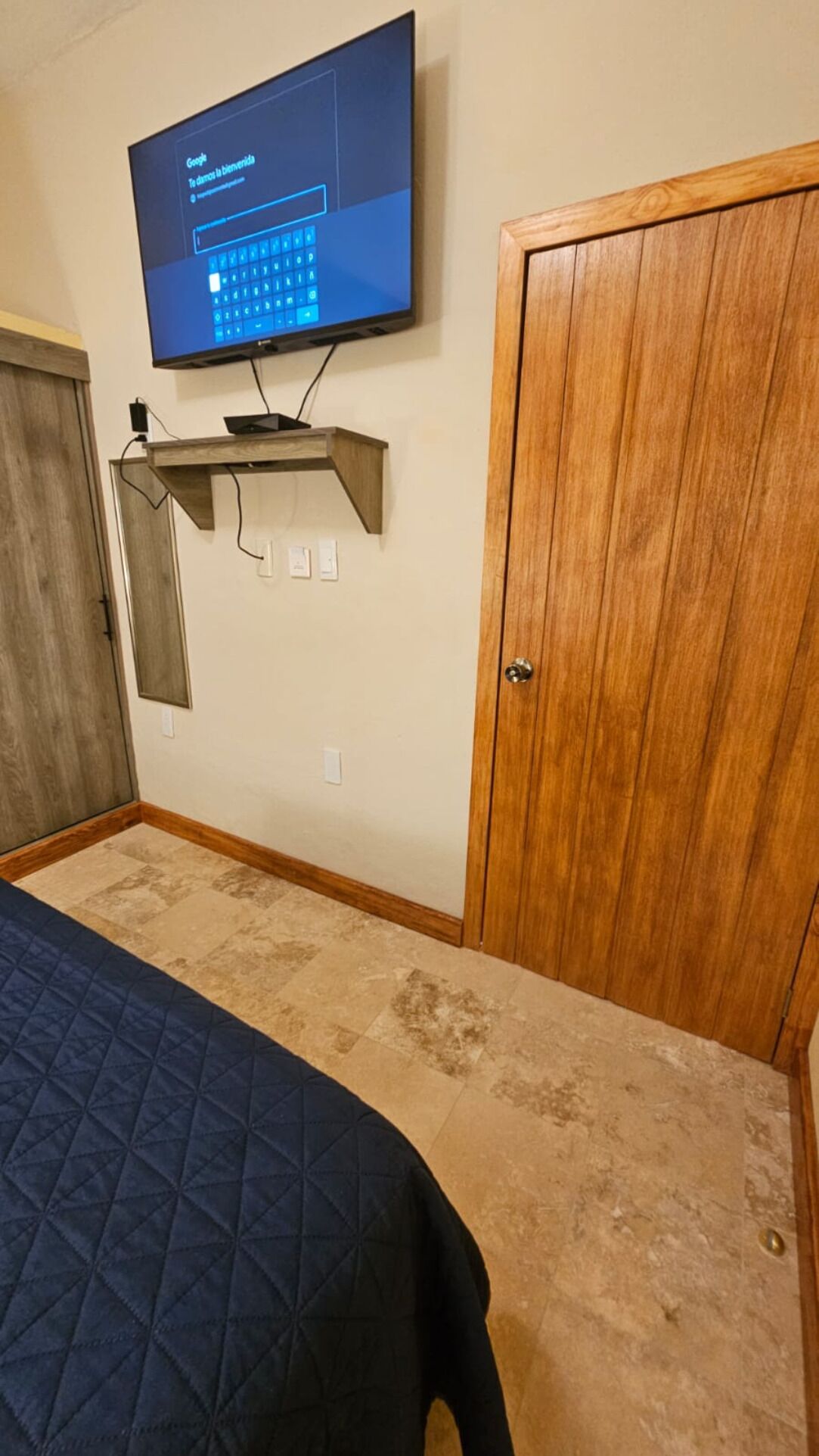 A bedroom with a bed, a television mounted on a wooden shelf, a wooden door, and a light switch on the wall.