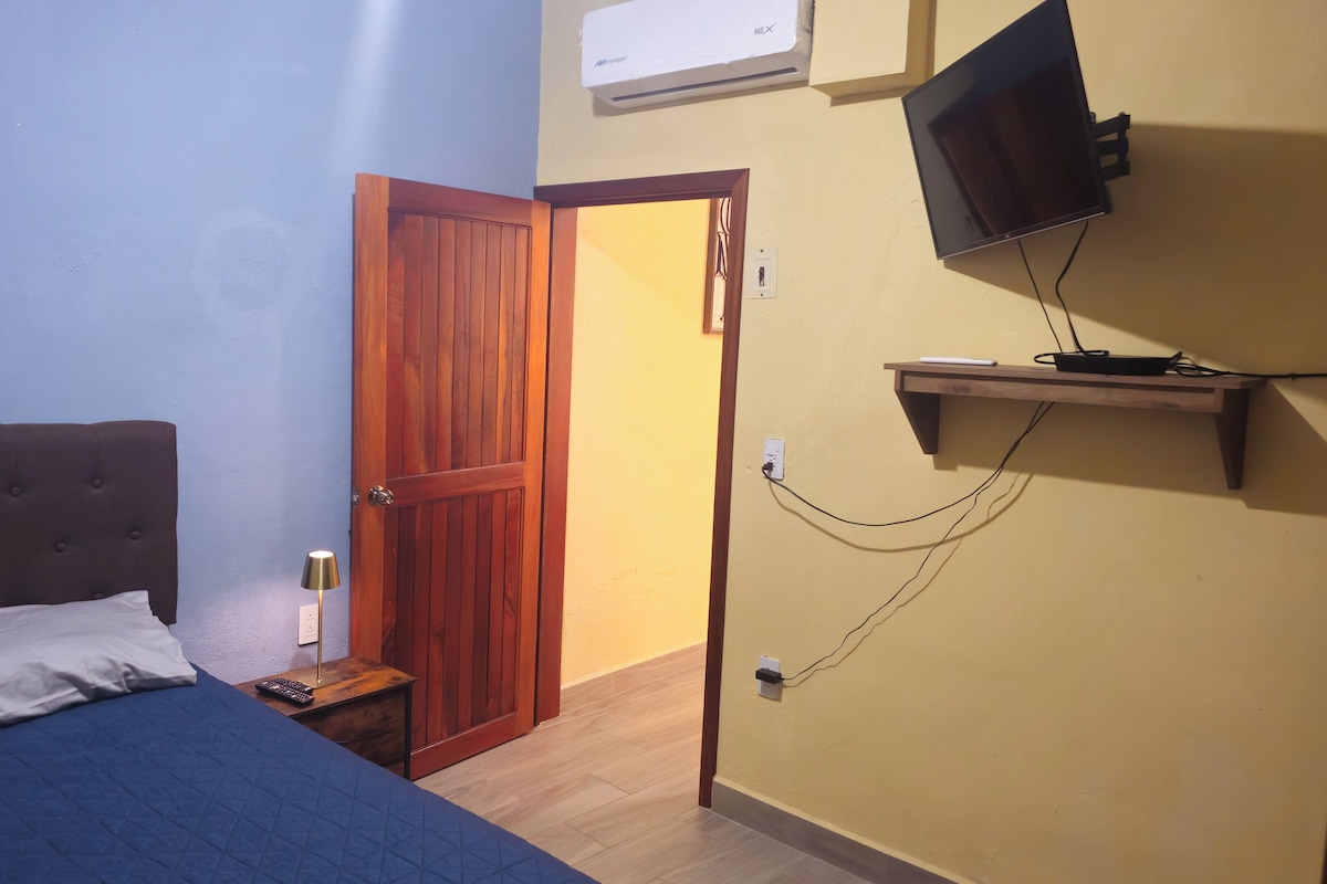 A bedroom with a bed, a nightstand, a lamp, a TV mounted on the wall, and a wooden door.