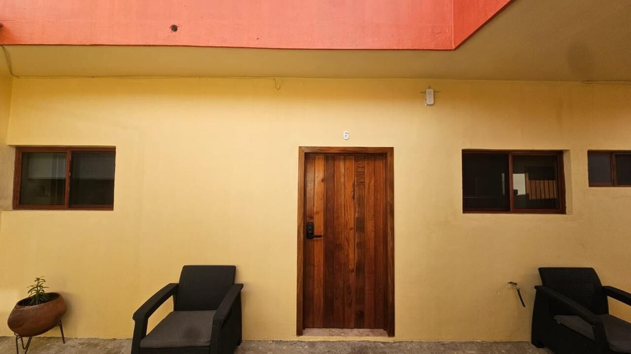 Un edificio con una puerta de madera marcada con el número 6, dos sillas negras y una planta en maceta.