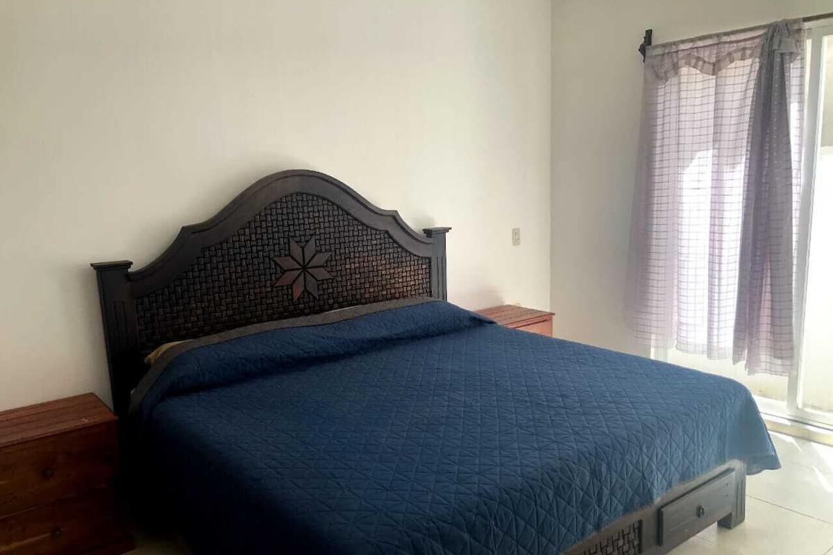 A bed with a dark headboard and a blue quilted bedspread in a room with a tiled floor and a window with curtains.