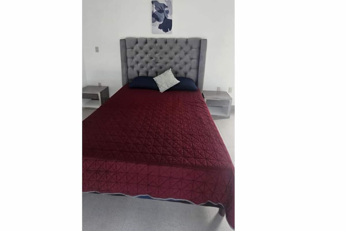 A bedroom with a tufted headboard, a red quilted bedspread, a white pillow, and a small bedside table with a lamp.
