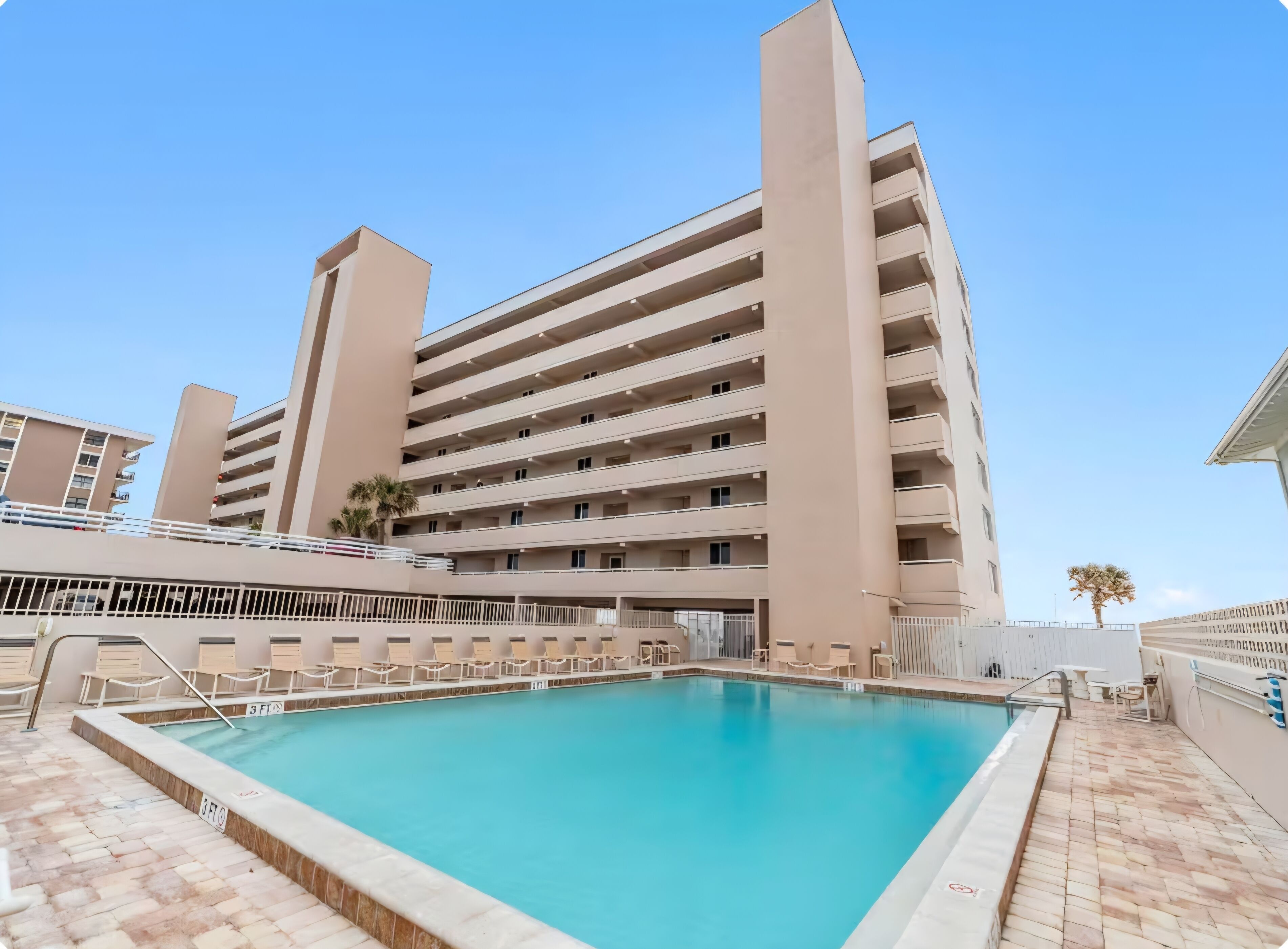 Un hotel de varios pisos con piscina, mobiliario exterior y cielo despejado.