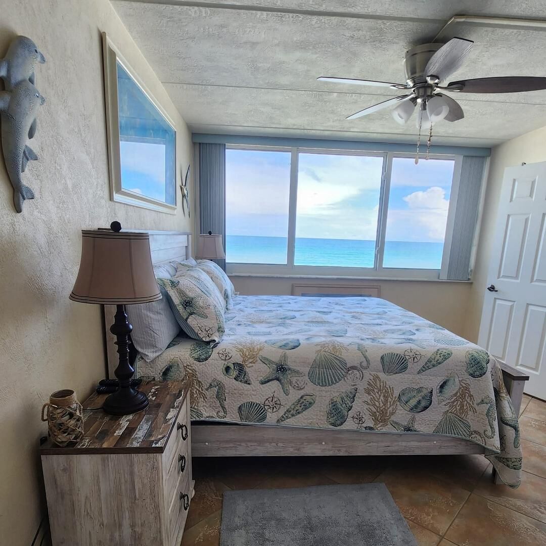 A bedroom with a bed, a ceiling fan, a window with a view of the ocean, and a lamp on a nightstand.