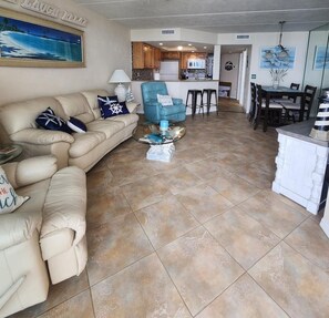 A living room with a beige sofa, a blue armchair, and a glass coffee table. A kitchen is visible in the background with wooden cabinets and a white refrigerator. - Paradise in the Hidden Gem Ponce Inlet, professionally, beautifully decorated, (Ponce Inlet)