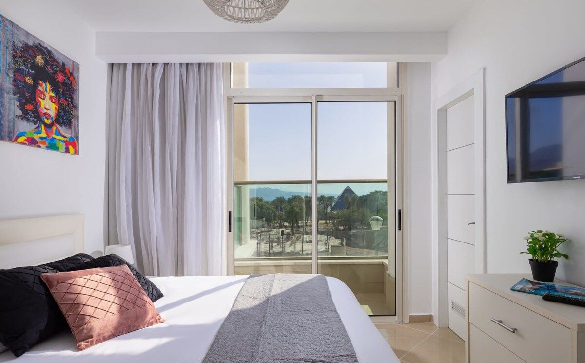 A modern bedroom with a large bed, a sliding glass door to a balcony, a mounted TV, and a painting on the wall.
