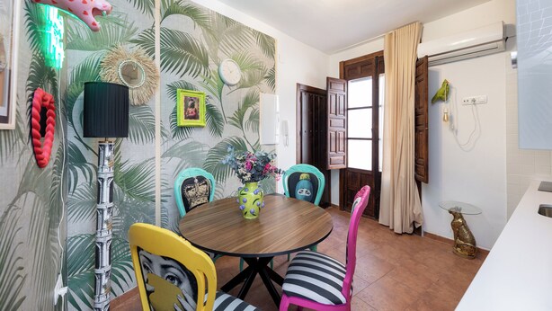 A dining area with a round wooden table, colorful chairs, a decorative wall with palm leaf patterns, a wall-mounted phone, and a small table with a glass top. - The Crown (1stRight) (Granada)