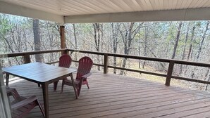 A covered outdoor deck with a table and chairs, surrounded by a railing, overlooking a forested area.