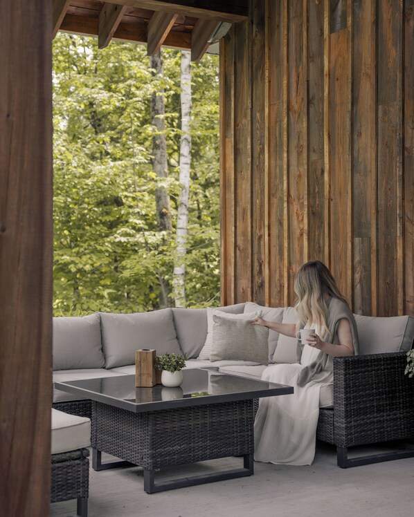 Un espace extérieur aménagé, abrité, avec un canapé et des meubles en osier, une petite table agrémentée d’une plante, et une vue sur les arbres à travers un mur en bois.