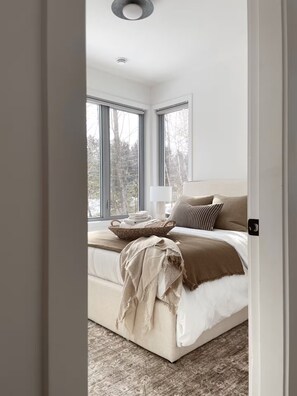 Une chambre à coucher avec un grand lit, une table de chevet, une lampe et une vue sur les arbres par la fenêtre.