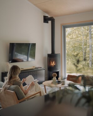 Un salon moderne doté d’une cheminée, d’un téléviseur fixé au mur et d’une grande fenêtre donnant sur la forêt.