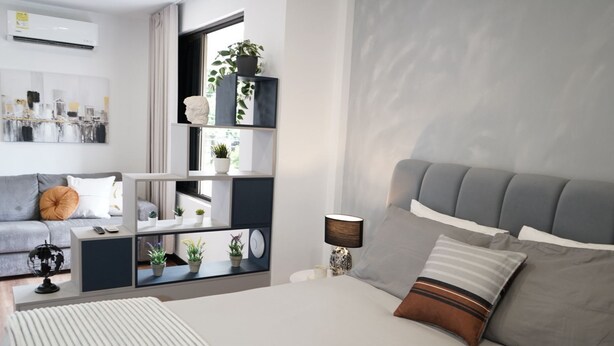 Une chambre moderne avec un lit recouvert d’un tissu gris, une table de chevet avec une lampe, un canapé gris et une bibliothèque intégrée agrémentée de plantes.