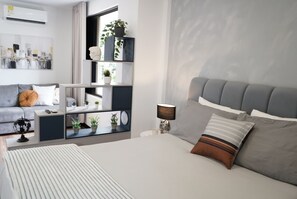 A modern bedroom with a grey upholstered bed, a bedside table with a lamp, a grey sofa, and a built-in shelving unit with plants. - Cozy apartment in central Limonar with air conditioning (Cali)