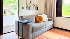 Un salon moderne avec un canapé gris, une petite table d’appoint sur laquelle se trouve une boisson, et une grande fenêtre avec des rideaux.