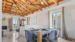 A spacious dining area with a wooden ceiling, a modern table with chairs, and a kitchen in the background. - Villa Kallia (Spetses)