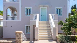 A two-story building with a light pink facade, a central entrance with a blue door, and a staircase leading to the upper floor. - Villa Kallia (Spetses)