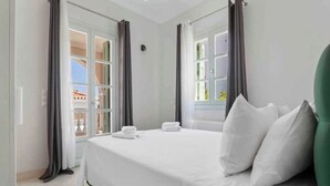 A hotel room with a large bed, two windows with curtains, and a view of the outside. - Villa Kallia (Spetses)