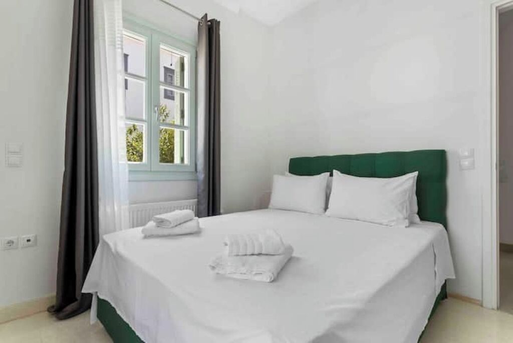 A neatly made bed with white linens and a green headboard in a bright room with a window and curtains.