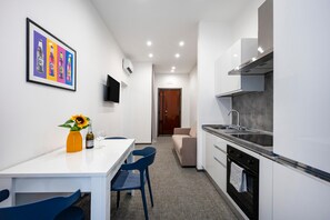 Een moderne keuken met witte kasten, een roestvrijstalen spoelbak en een inbouwoven. Er is een eethoek met een witte tafel en blauwe stoelen. Een ingelijste afbeelding hangt aan de muur, en op de tafel staat een vaas met een zonnebloem.
