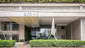 Un edificio moderno con un cartello 'D/ONE', un caffè con il cartello 'SUCAS SALADAS + CAFES + SANDWICHES' e un siepe curata di fronte.