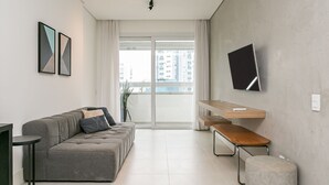 Un soggiorno moderno con un divano grigio, una panca in legno, un poggiapiedi rotondo, una TV a schermo piatto montata a parete e un'ampia finestra con vista sugli edifici.