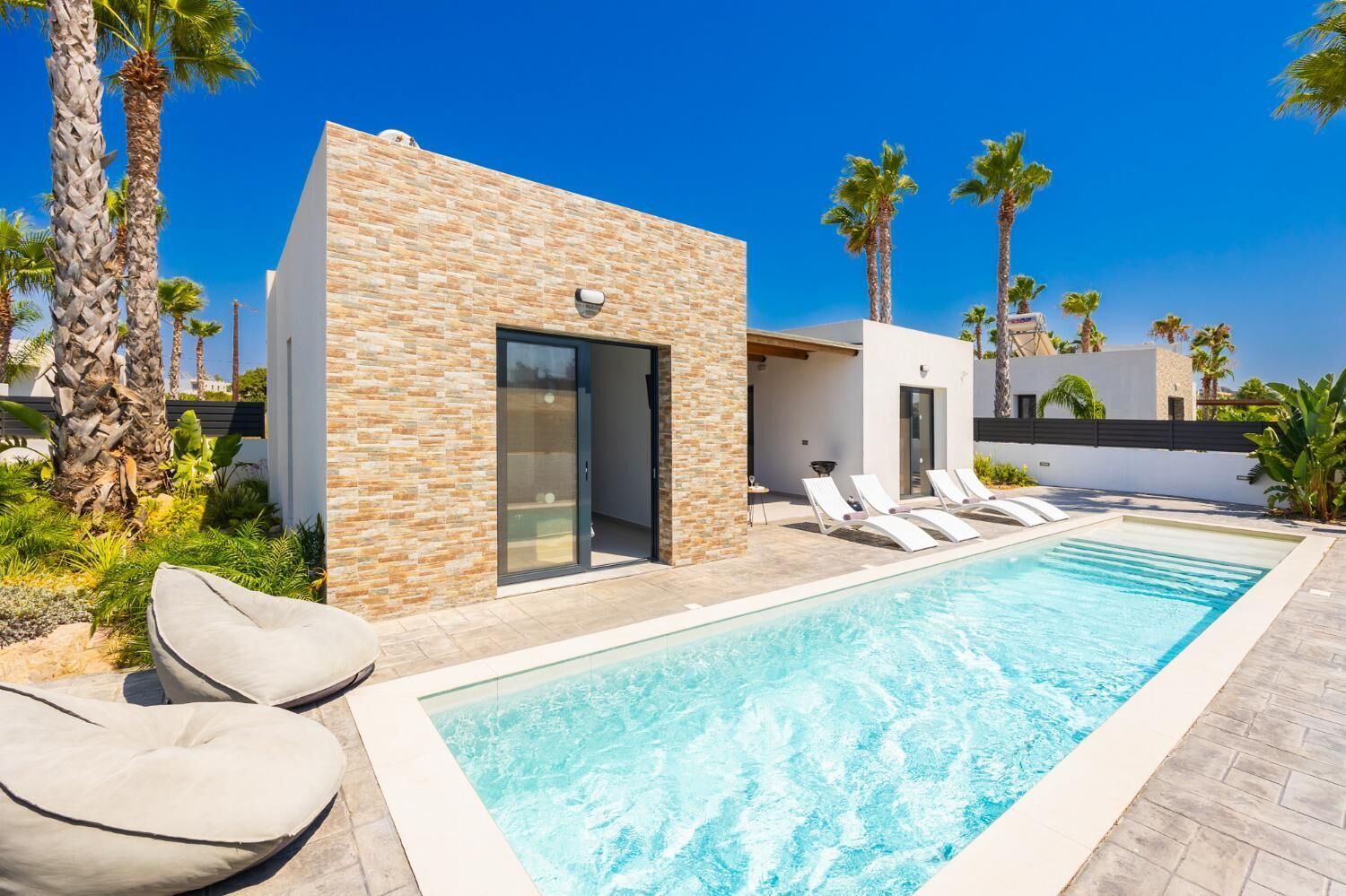 Ein moderner Poolbereich mit rechteckigem Pool, Liegestühlen und einem Gebäude aus Stein mit Glastür.