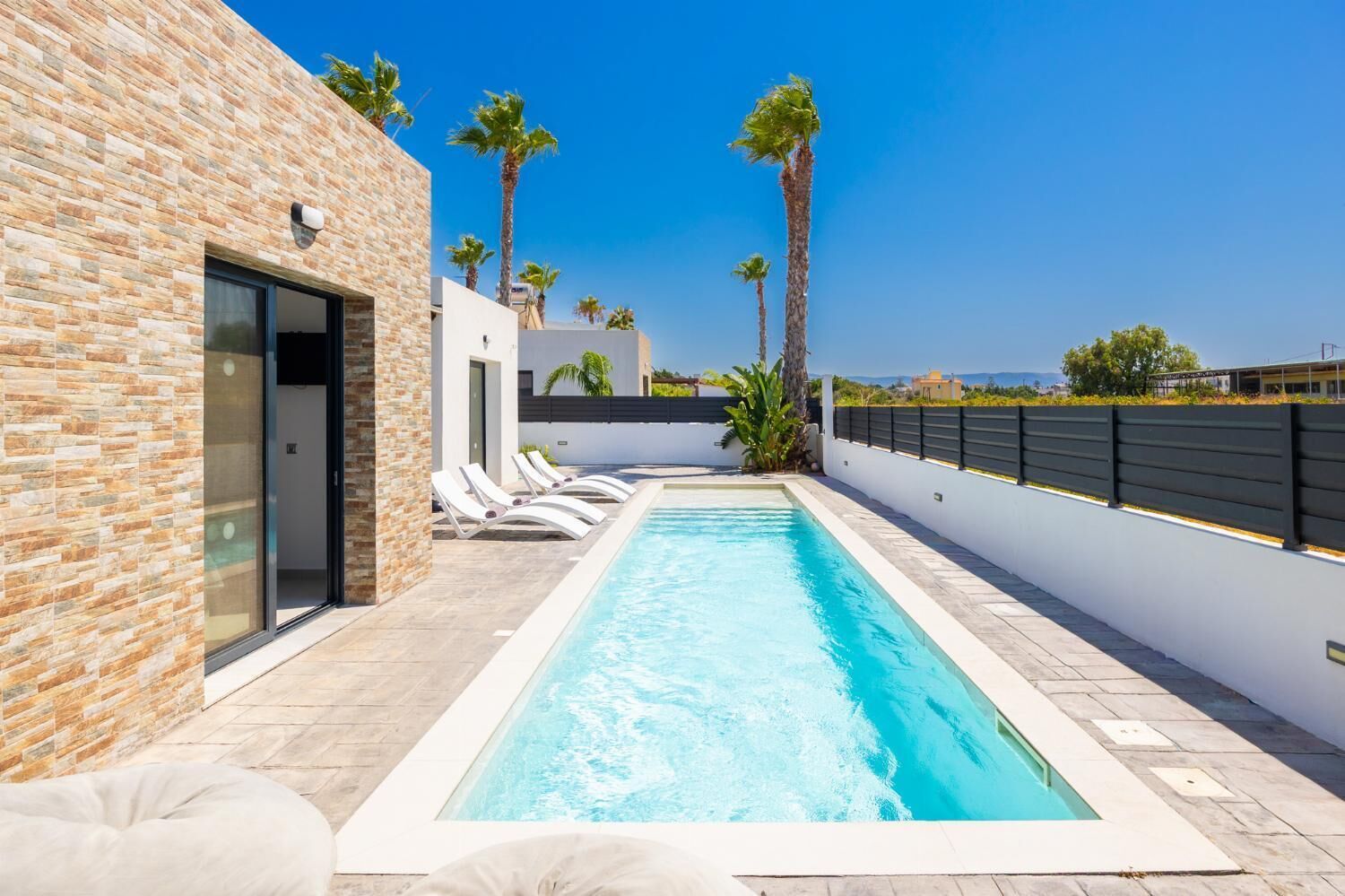 A swimming pool with lounge chairs, a stone wall, and palm trees.