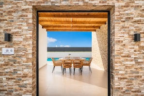 A covered outdoor dining area with a pool view, a wooden pergola, and a stone wall.